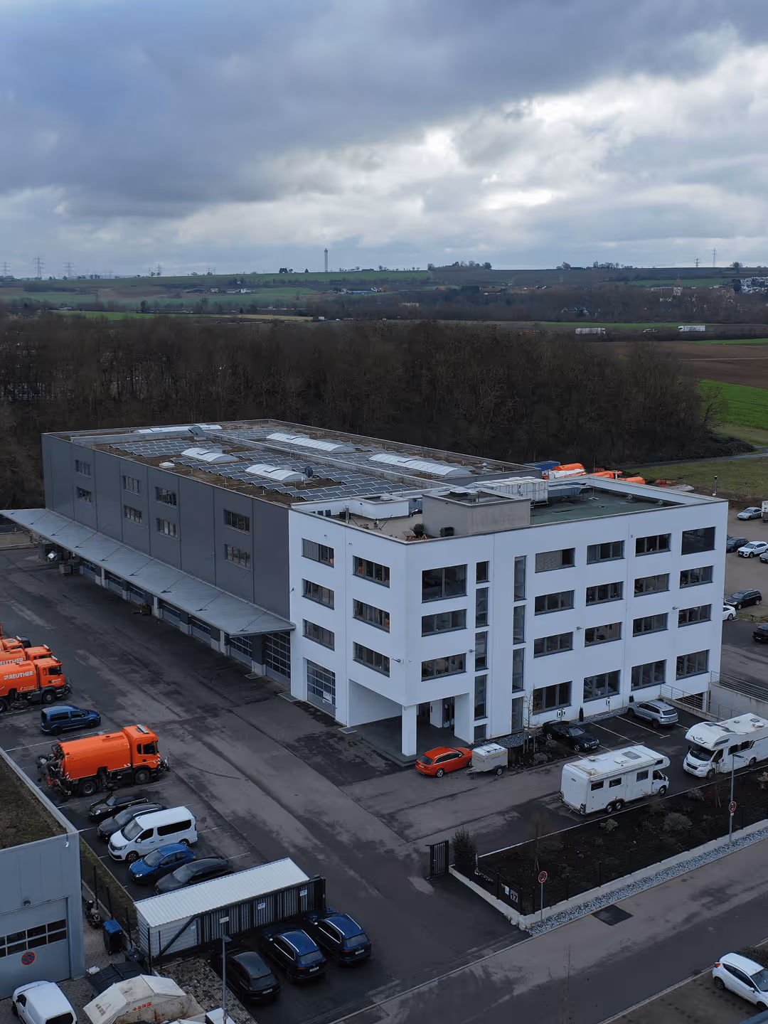 Modernes, mehrstöckiges Bürogebäude mit grauer und weißer Fassade, umgeben von geparkten Fahrzeugen unter bewölktem Himmel.