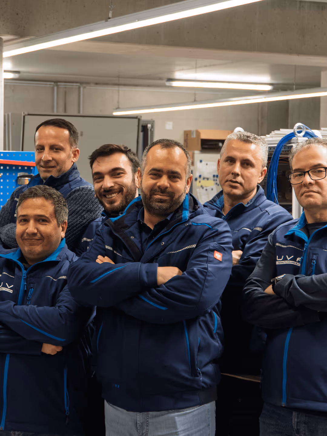 Sechs Männer in blauen Jacken stehen in Industrieraum, lächeln mit verschränkten Armen.