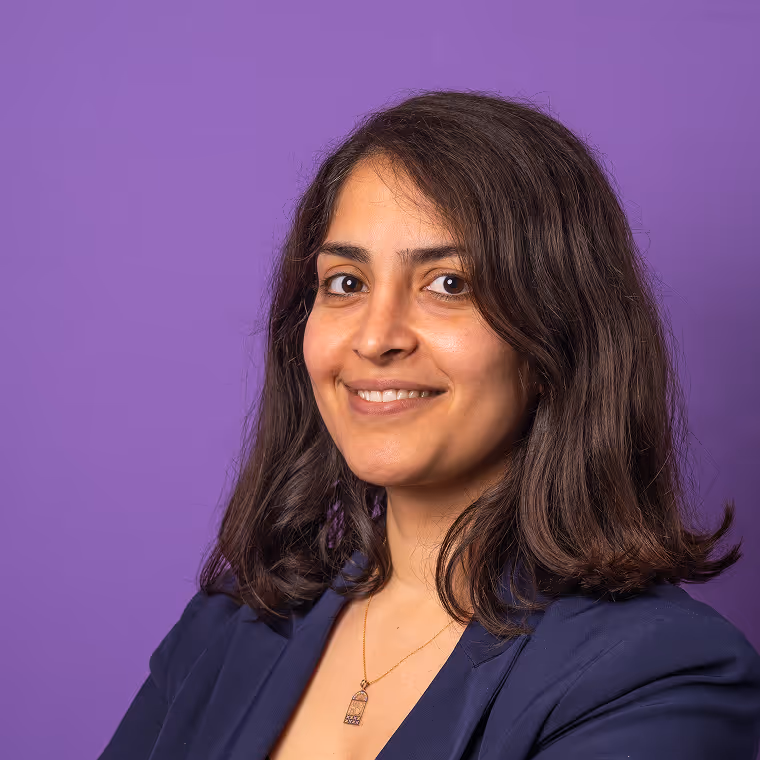 Smiling woman with shoulder-length dark hair wearing a navy blazer and pendant necklace against a purple background.