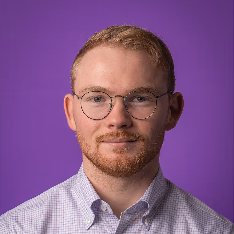 Portrait of a young man with light brown hair, short beard, round glasses, and a checkered shirt against a purple background.