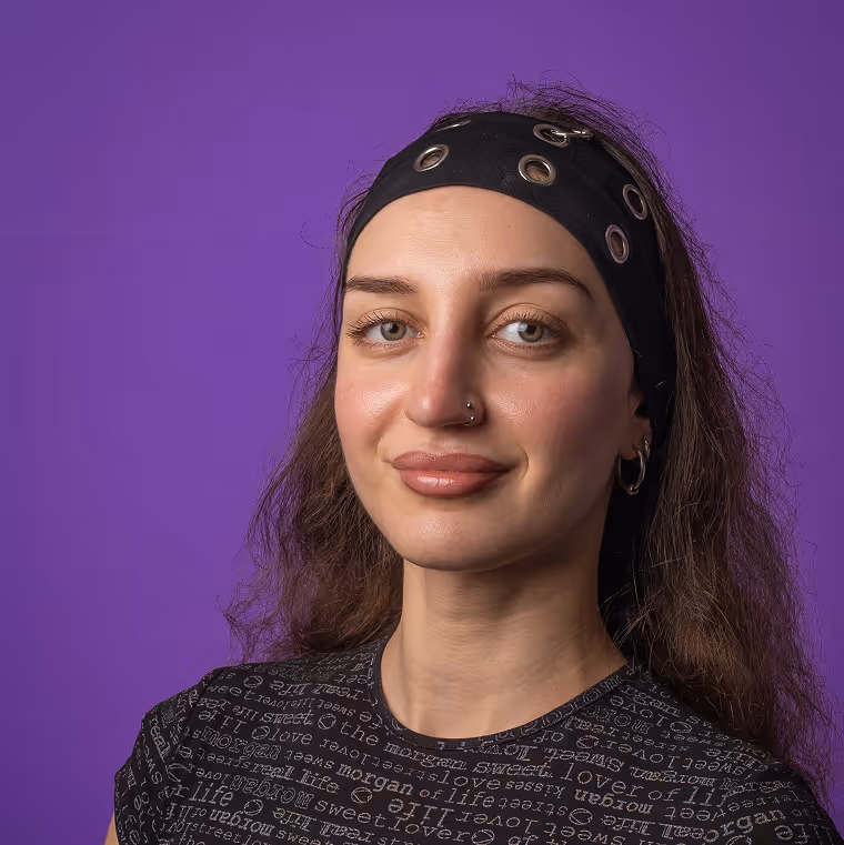 Young woman with a black headband and nose piercing smiling against a purple background.
