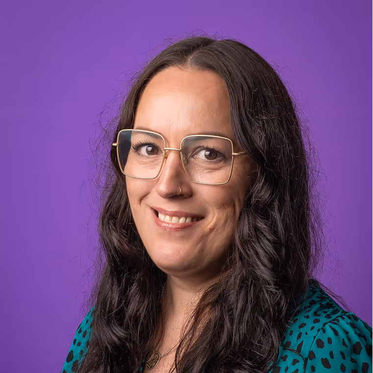 Smiling woman with long dark hair, gold-rimmed square glasses, and a nose ring, wearing a teal top with black spots against a purple background.