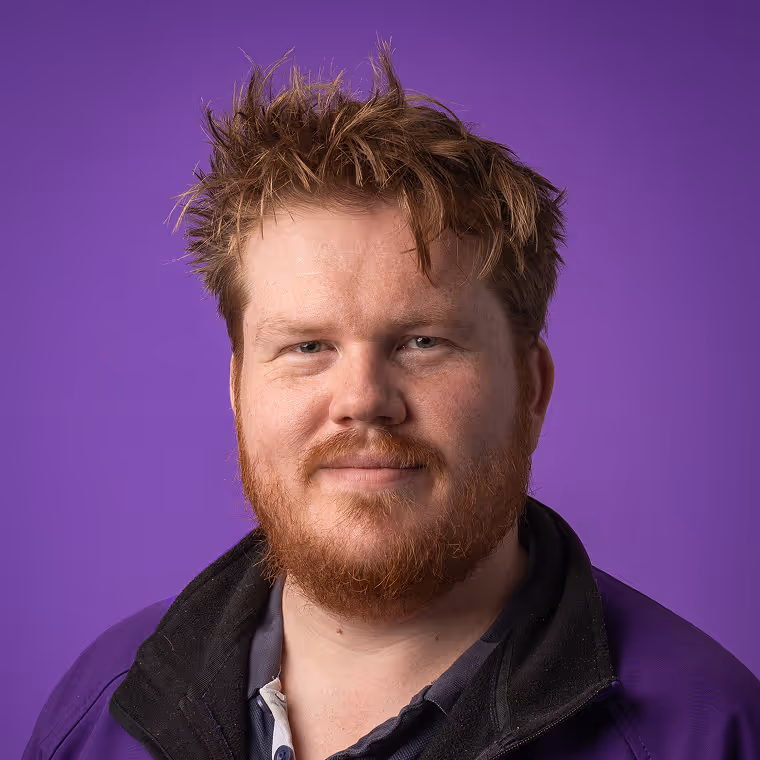 Man with messy reddish hair and beard wearing a purple jacket against a purple background.