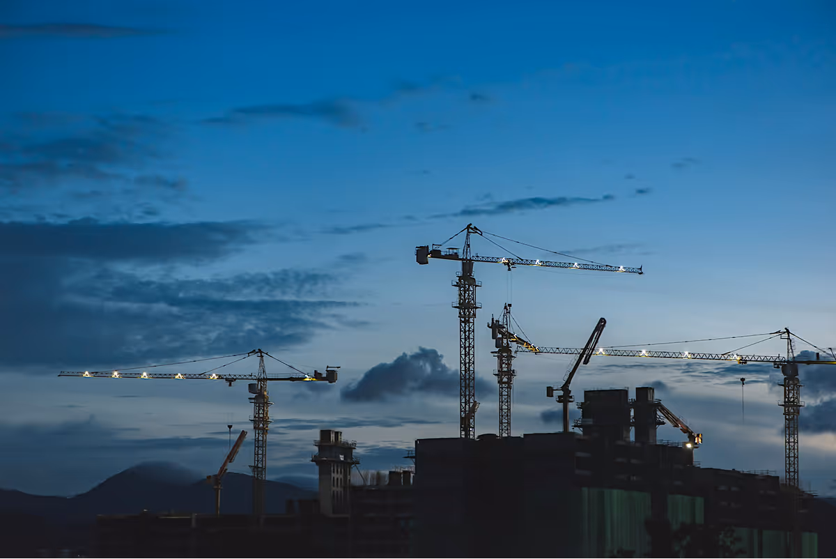 Construction cranes illuminated at dusk above building structures with a mountain silhouette in the background.