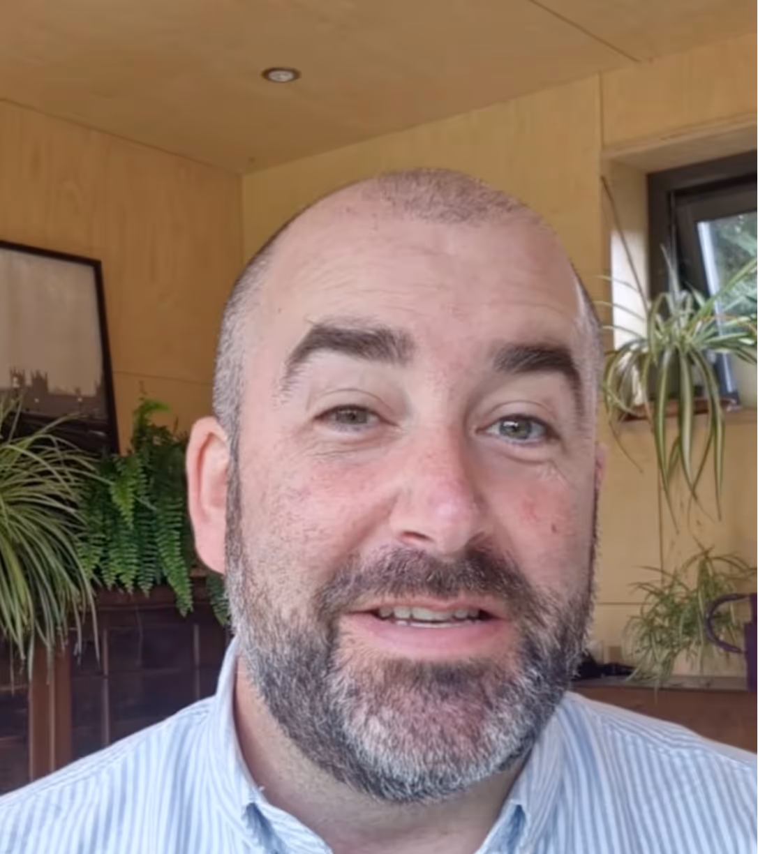 Smiling man with a beard and short hair indoors, with plants and wooden walls in the background.