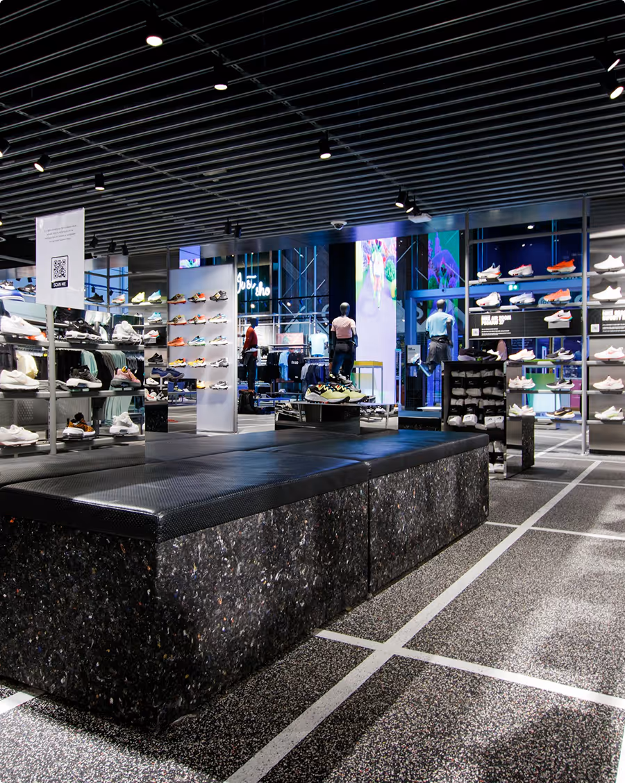 Modern sneaker store interior with black seating bench, illuminated shelves displaying various athletic shoes, and mannequins dressed in sportswear.