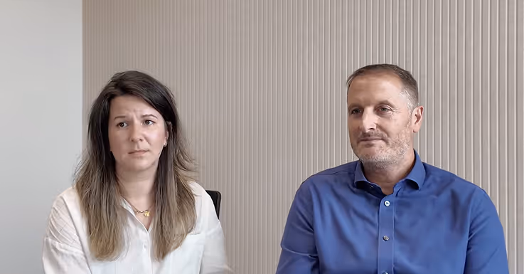 Man and woman sitting side by side against a striped beige wall, both looking forward.