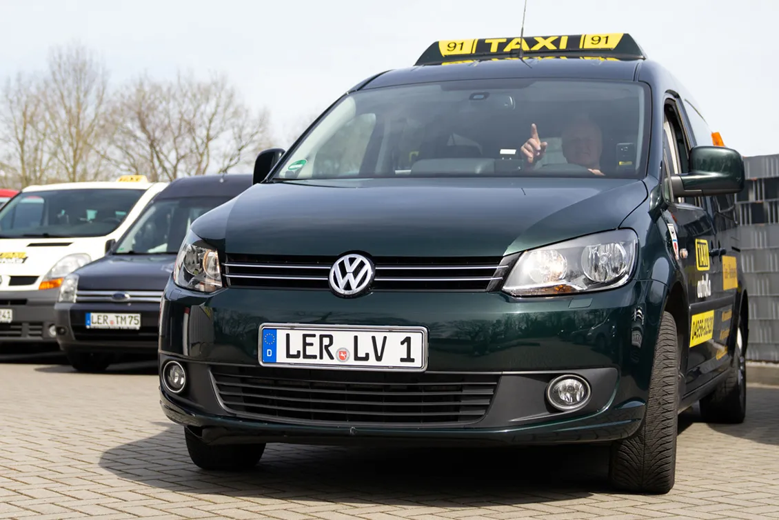 Dunkelgrünes Volkswagen-Taxi mit eingeschaltetem Scheinwerfer und Taxischild auf dem Dach, Fahrer hebt Finger, weitere Taxis im Hintergrund.