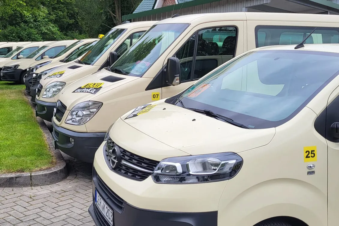 Reihe von cremefarbenen Taxi-Vans, geparkt auf einem gepflasterten Parkplatz neben einem Rasen.