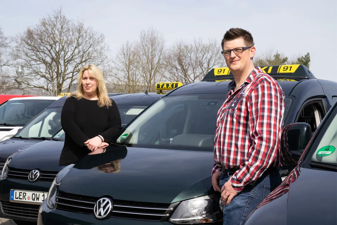 Zwei Personen stehen vor mehreren schwarzen Volkswagen-Taxis mit gelben Aufbauen, die Nummer 91 zeigen, an einem sonnigen Tag.