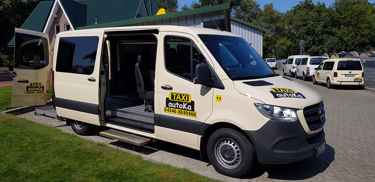 Beige Mercedes-Taxi-Van mit geöffneten Türen, geparkt auf einem gepflasterten Bereich neben einem grünen Rasen und mehreren weiteren Taxis im Hintergrund.