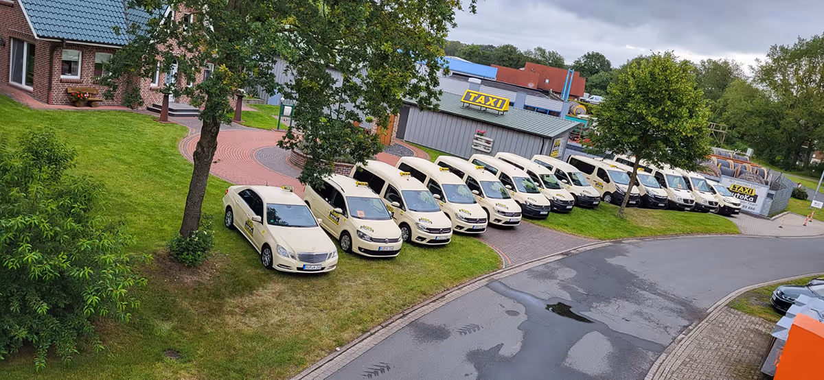 Mehrere weiße Taxis und Kleinbusse parken auf einem grasbewachsenen Bereich neben einer Straße vor einem Gebäude mit einem gelben TAXI-Schild.