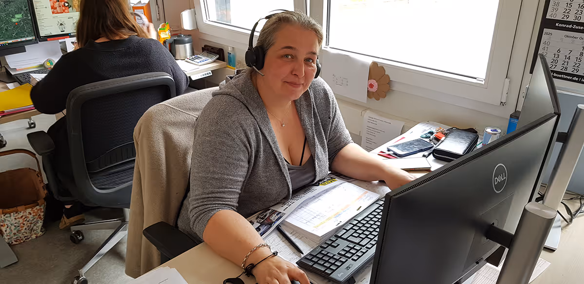 Eine Frau mit Headset sitzt an einem Schreibtisch vor zwei Monitoren und lächelt in die Kamera.