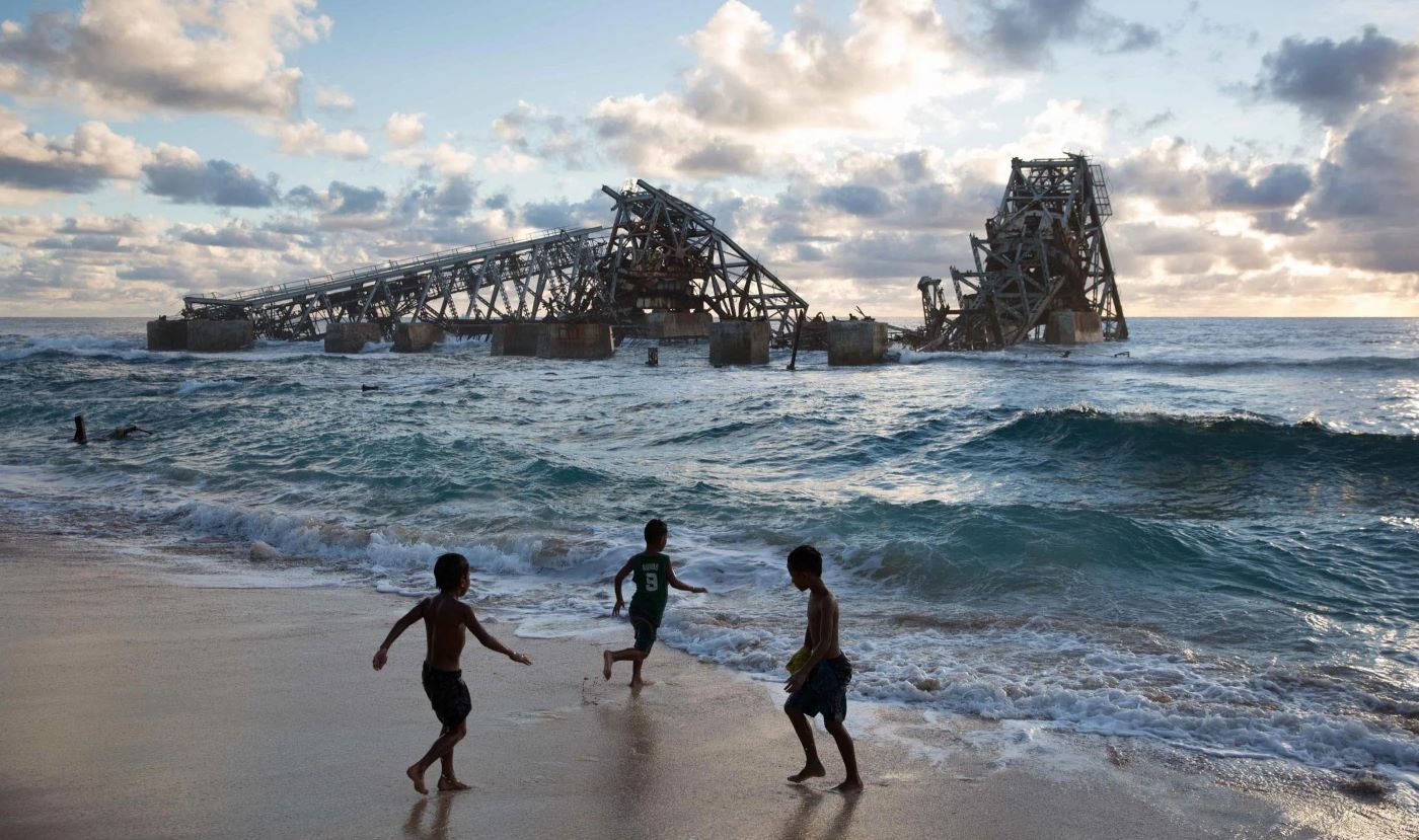 Three Nauruan children play on the beach in front of the collapsed phosphate loading cantilever, now half-submerged in the Pacific off Aiwo district.