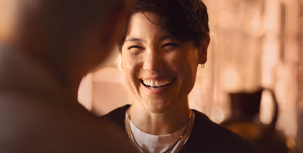 A person smiling warmly during a conversation, wearing layered necklaces and a black top, with soft lighting creating a cozy atmosphere.