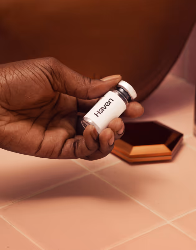 A hand holding a Haven Health product on a pink bathroom counter, surrounded by makeup items and a leather pouch.