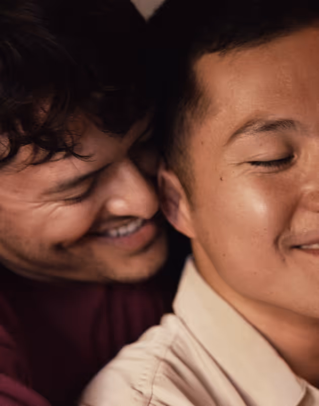 Close-up of a joyful couple embracing; one man smiles as he leans in toward the other, who is also smiling with his eyes closed.