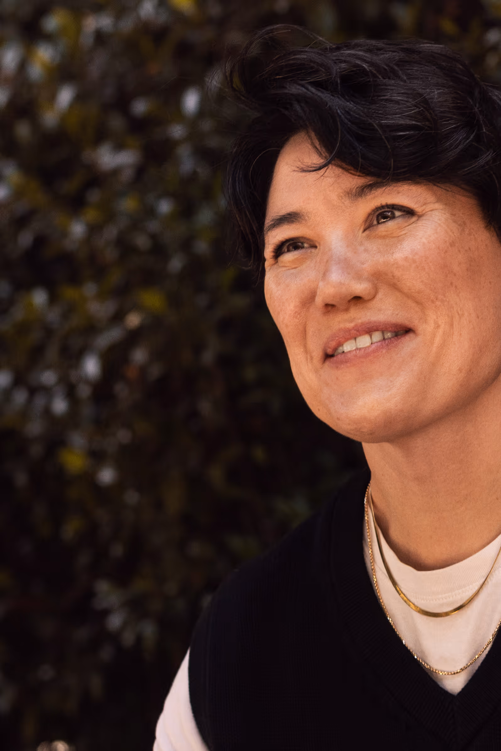 Person smiling outdoors in front of a leafy background, wearing layered gold necklaces, a white shirt, and a black vest.