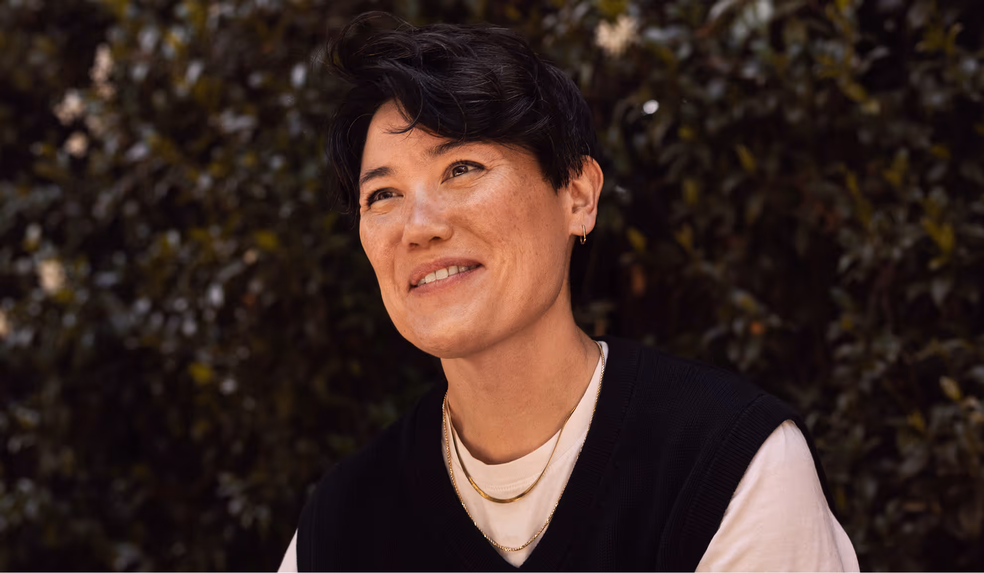 Person smiling outdoors in front of a leafy background, wearing layered gold necklaces, a white shirt, and a black vest.