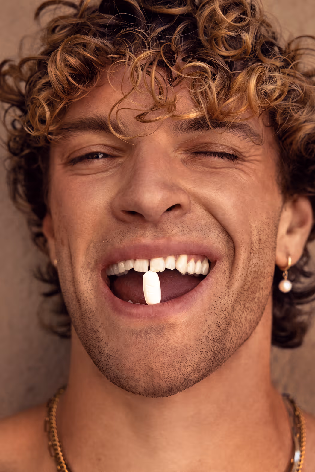 Close-up of man with curly hair holding a white pill between his teeth and winking.