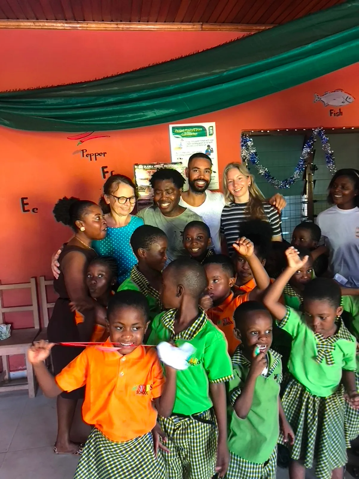 Eine Gruppe von Erwachsenen und Kindern posiert in einem bunt dekorierten Klassenzimmer; vorne tragen die Kinder grüne und orange Schuluniformen, während die Erwachsenen im Hintergrund zusammenstehen und lächeln.