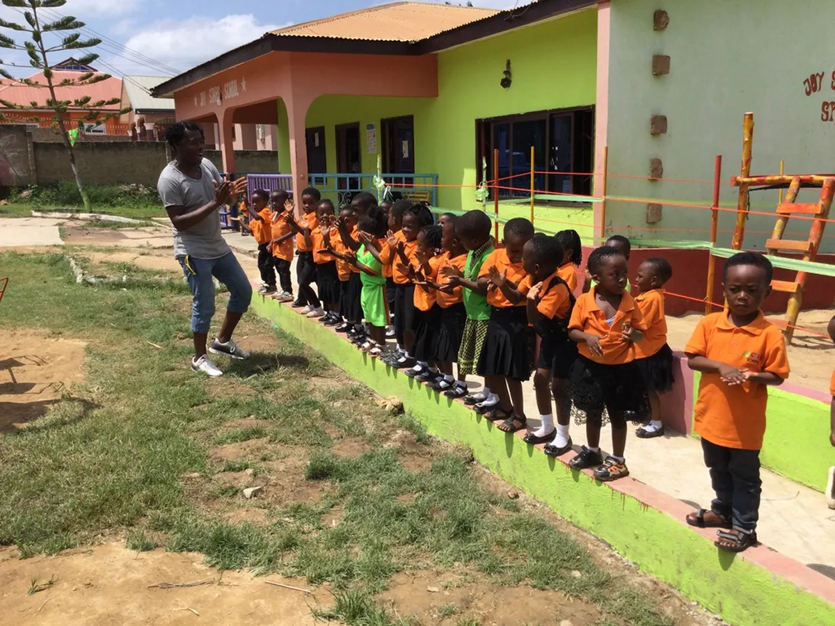 Eine Gruppe kleiner Kinder in orangen Schuluniformen steht in einer Reihe auf einer niedrigen Mauer vor einem bunten Schulgebäude, während ein Erwachsener ihnen gegenübersteht und klatscht.