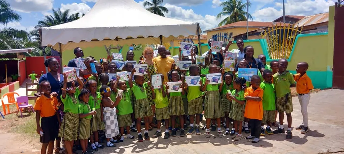 Eine große Gruppe von Schulkindern in grünen und orangen Uniformen steht gemeinsam mit Erwachsenen vor einem Pavillon im Freien und hält stolz Urkunden und Hefte in die Höhe.