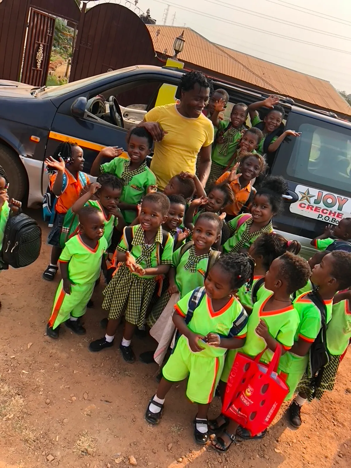 Eine große Gruppe von Kindern in grünen und karierten Schuluniformen steht fröhlich vor einem Kleinbus mit der Aufschrift Joy Stars, begleitet von einem Erwachsenen im gelben T-Shirt.