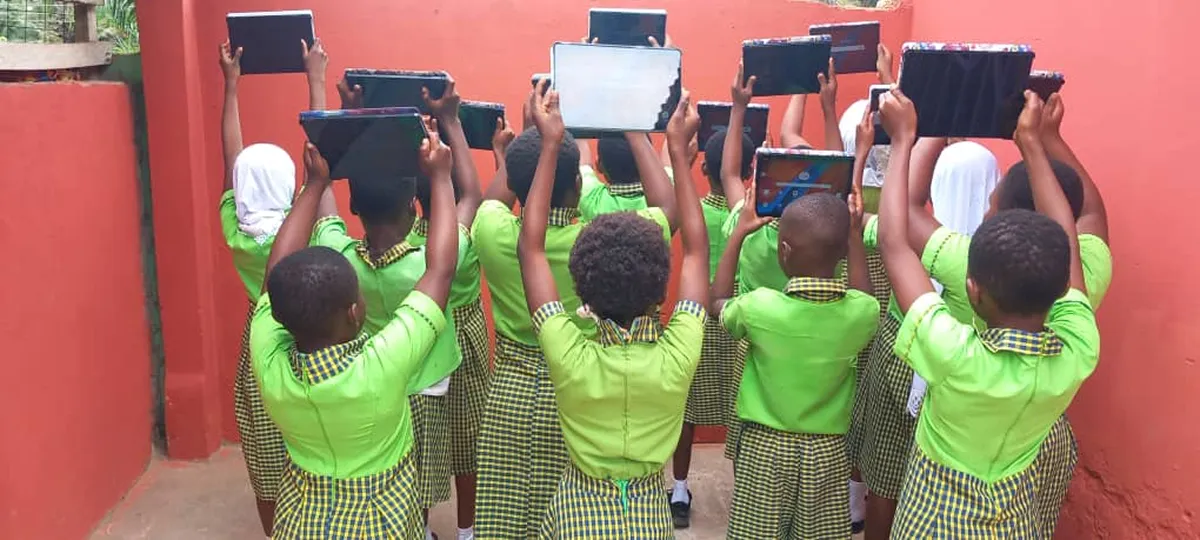 Kinder in grünen Schuluniformen halten Tablets über ihren Köpfen hoch, Rückenansicht vor einer roten Wand