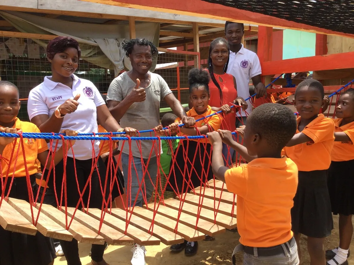 Schulkinder und Erwachsene auf dem Spielplatz, gemeinsam an einer bunten Holzbrücke mit roten Seilen