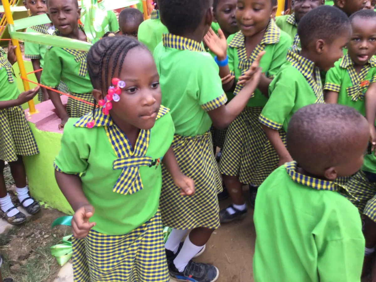 Nahaufnahme eines Mädchens in grüner und gelb karierter Schuluniform mit rosa Haarperlen, umgeben von anderen Kindern auf dem Schulhof in Ghana