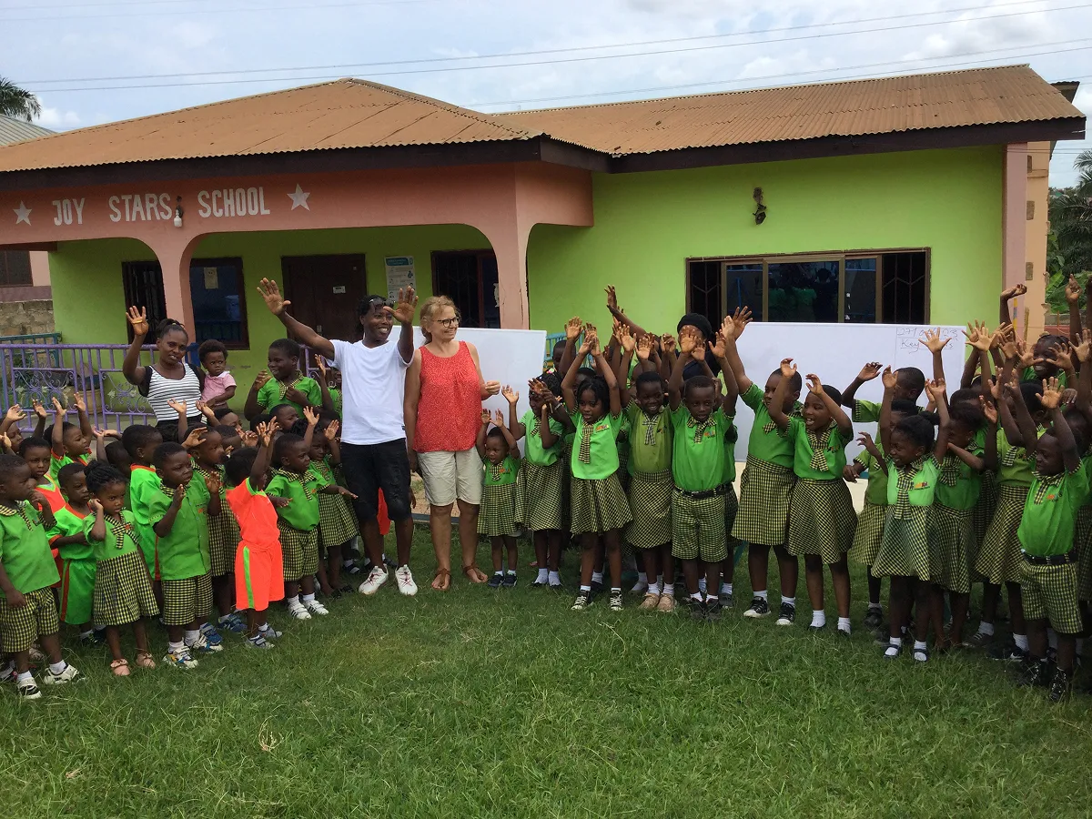 Großes Gruppenfoto von Kindern in grünen Schuluniformen mit zwei Erwachsenen vor der Joy Stars School in Ghana, alle heben fröhlich die Hände