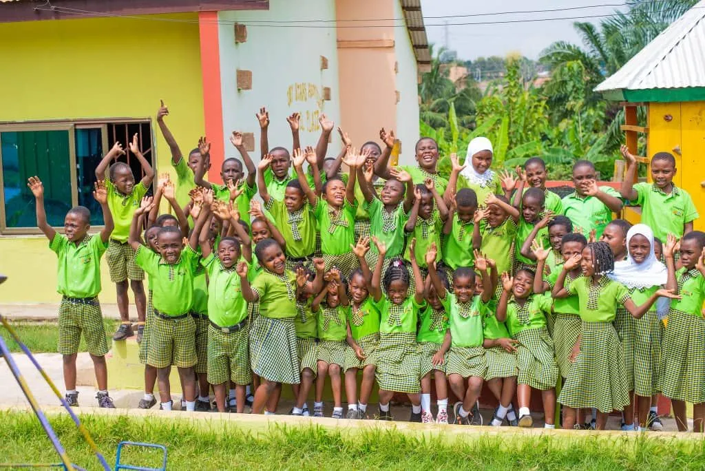 Große Gruppe von Schulkindern in grüner Uniform hebt fröhlich die Arme vor einem Gebäude in Ghana