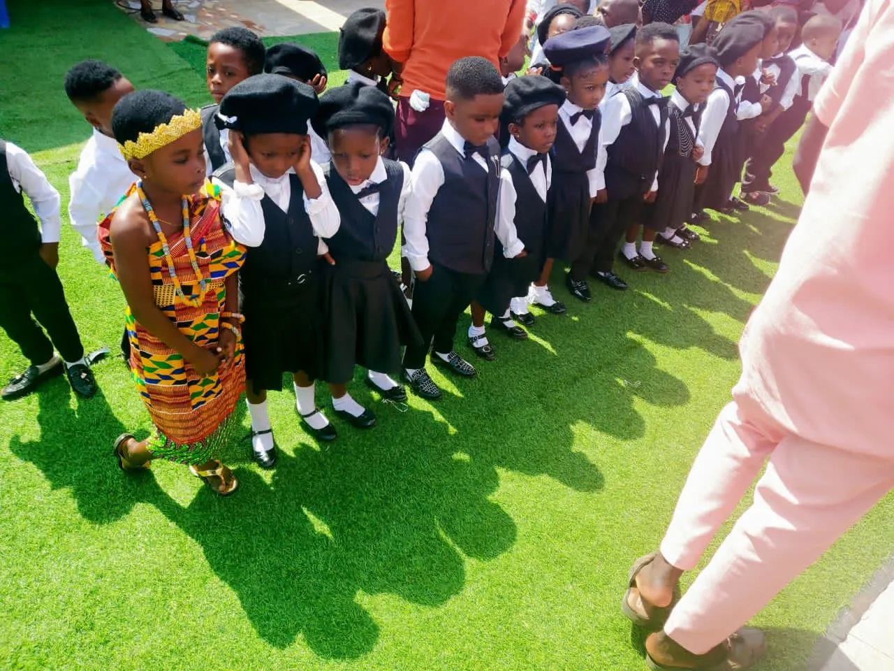 Kinder in Ghana in feierlicher Tracht und Schuluniform stehen in einer Reihe auf grünem Rasen