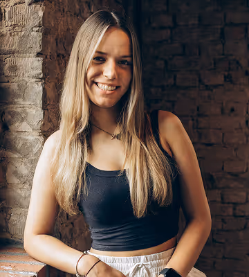 Junge Frau lächelt in heller Kleidung und sitzt vor einer Backsteinwand.