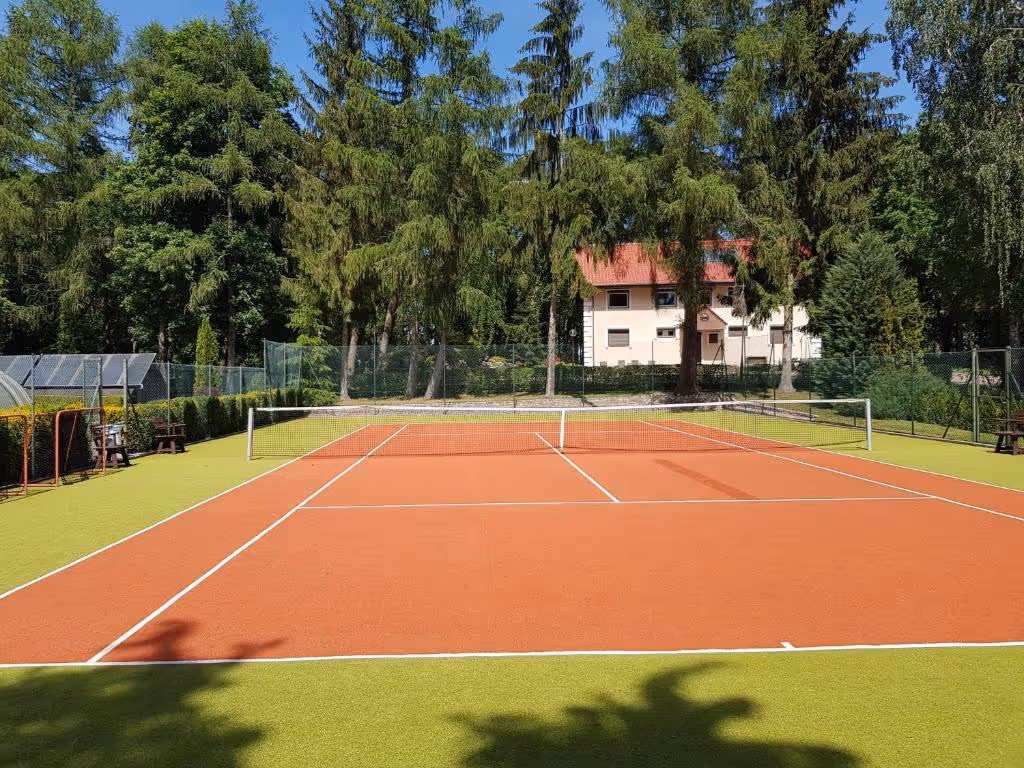 Tennisplatz mit rotem Belag und grünem Rand, umgeben von Bäumen und Gebäuden der Unterkunft.