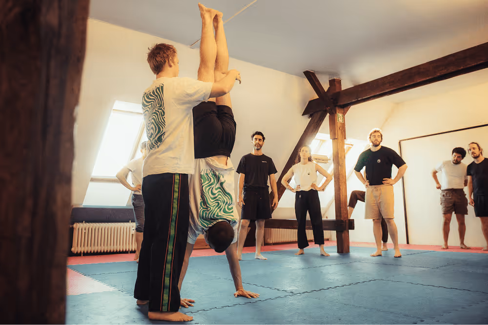 Trainer führt mit Teilnehmer eine Handstand Partnerübung während eines Movement Kurses vor und die Gruppe schaut zu.