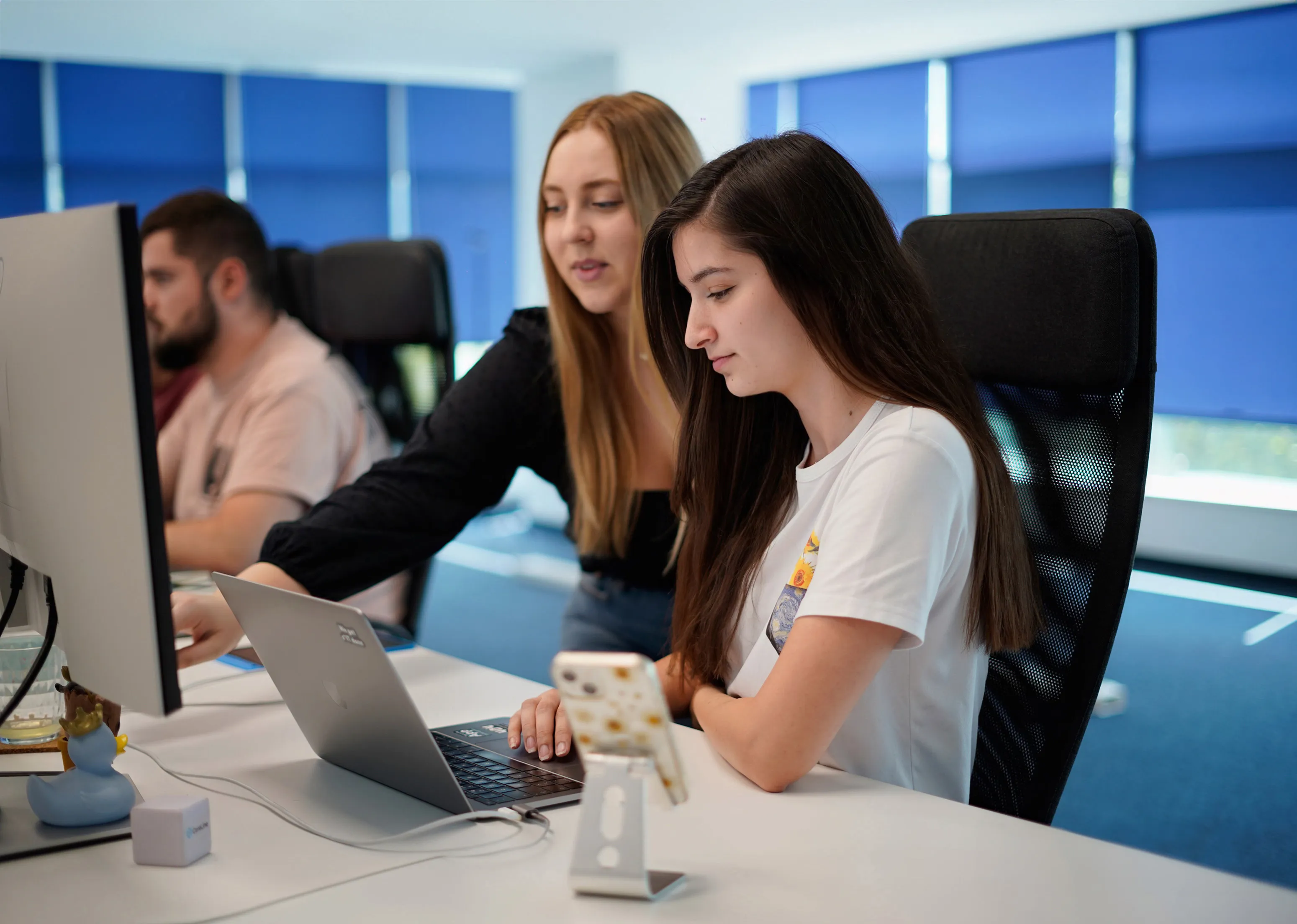 Two women working together at a desk with a laptop and desktop monitor in a modern office.