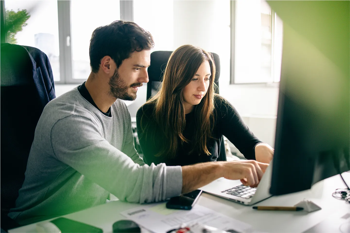 A man and woman working together at a desk, looking at a laptop screen and pointing at it in a bright office.