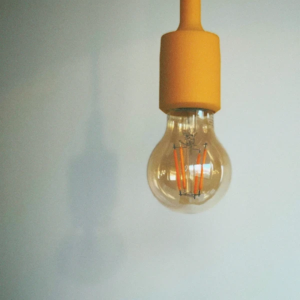 Clear glass light bulb with orange filaments hanging against a light gray wall.