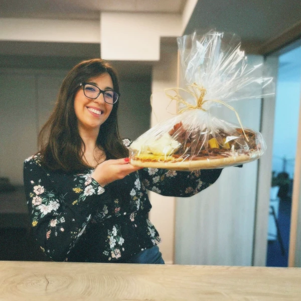 Smiling woman with glasses holding a wrapped gift basket tied with a bow, standing indoors.