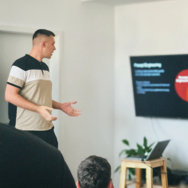 Man in a polo shirt giving a presentation to an audience with a laptop and a screen displaying graphics in the background.