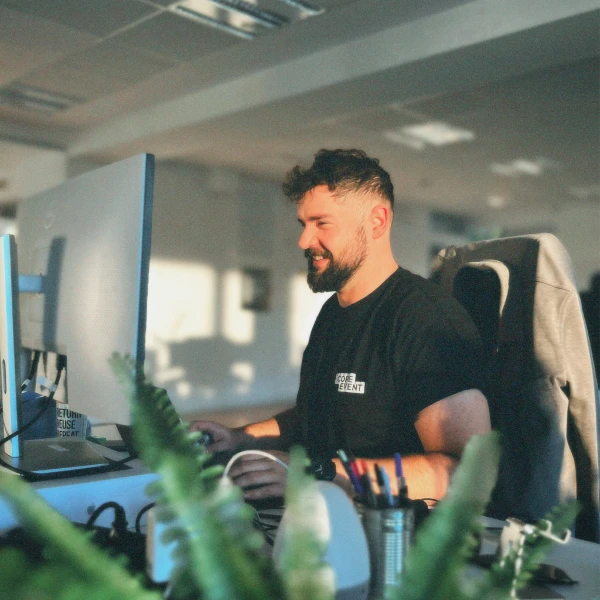 Bearded man smiling while working at a computer desk in a sunlit office.