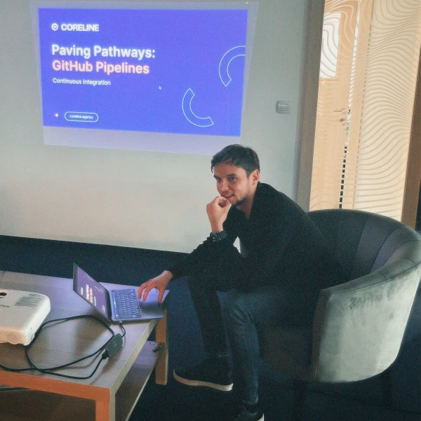 Man seated on a gray chair working on a laptop with a presentation titled 'Paving Pathways: GitHub Pipelines' projected on the wall.