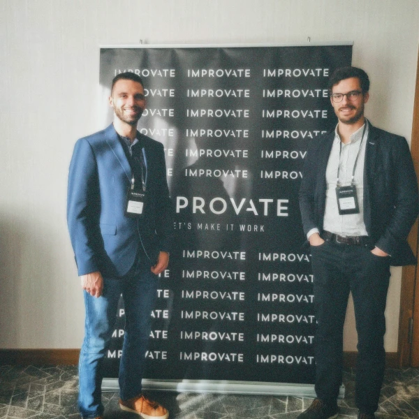 Two men in business attire standing on either side of a black banner with repeated 'IMPROVATE' text.