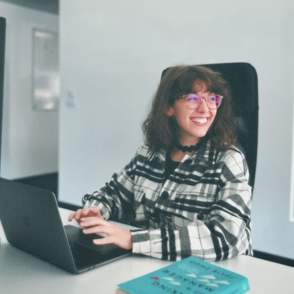 Smiling young woman with glasses and a black-and-white plaid jacket sitting at a table using a laptop, with a turquoise book in front of her.