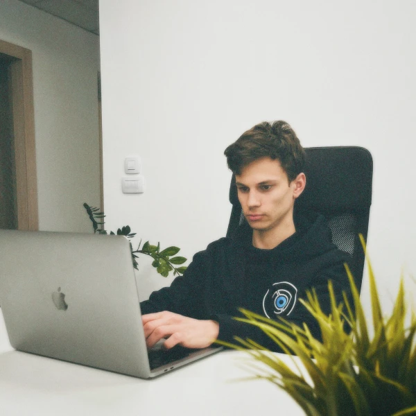 Young man focused on typing on a silver MacBook laptop at a white desk with plants in the background.