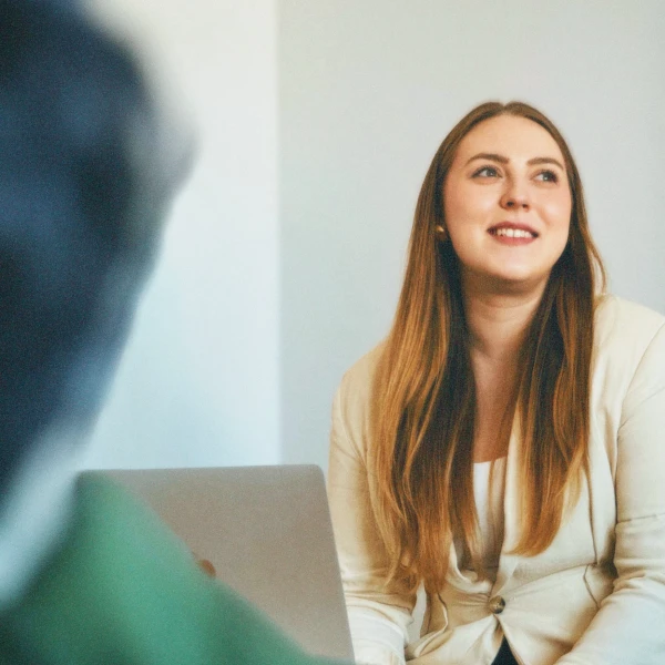 Smiling woman with long hair wearing a beige blazer engaged in a conversation indoors.