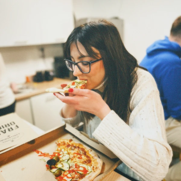 Woman with glasses and dark hair eating a slice of pizza in a kitchen setting.