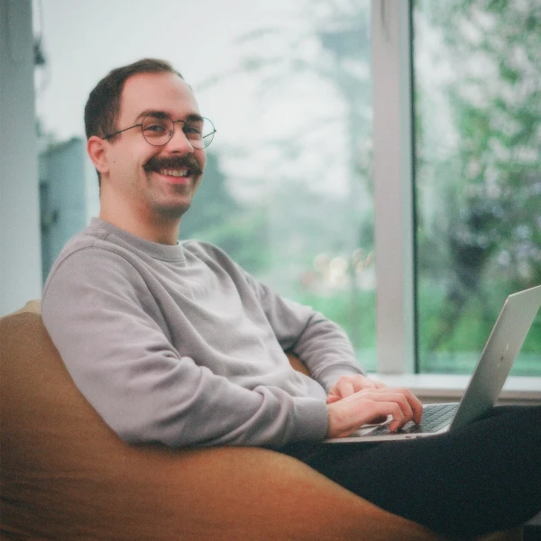 Smiling man with glasses and mustache sitting in a brown chair, typing on a laptop near a large window with green outdoor scenery.